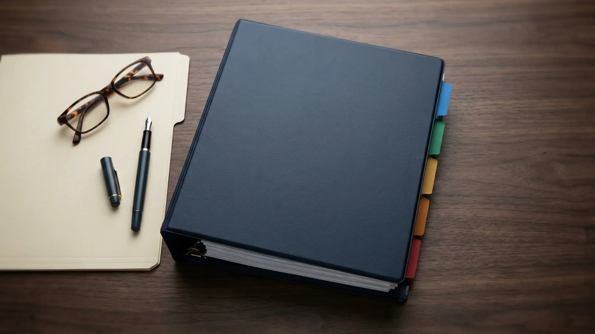 Closed navy mitigation binder with colored index tabs beside a file folder, fountain pen, and reading glasses